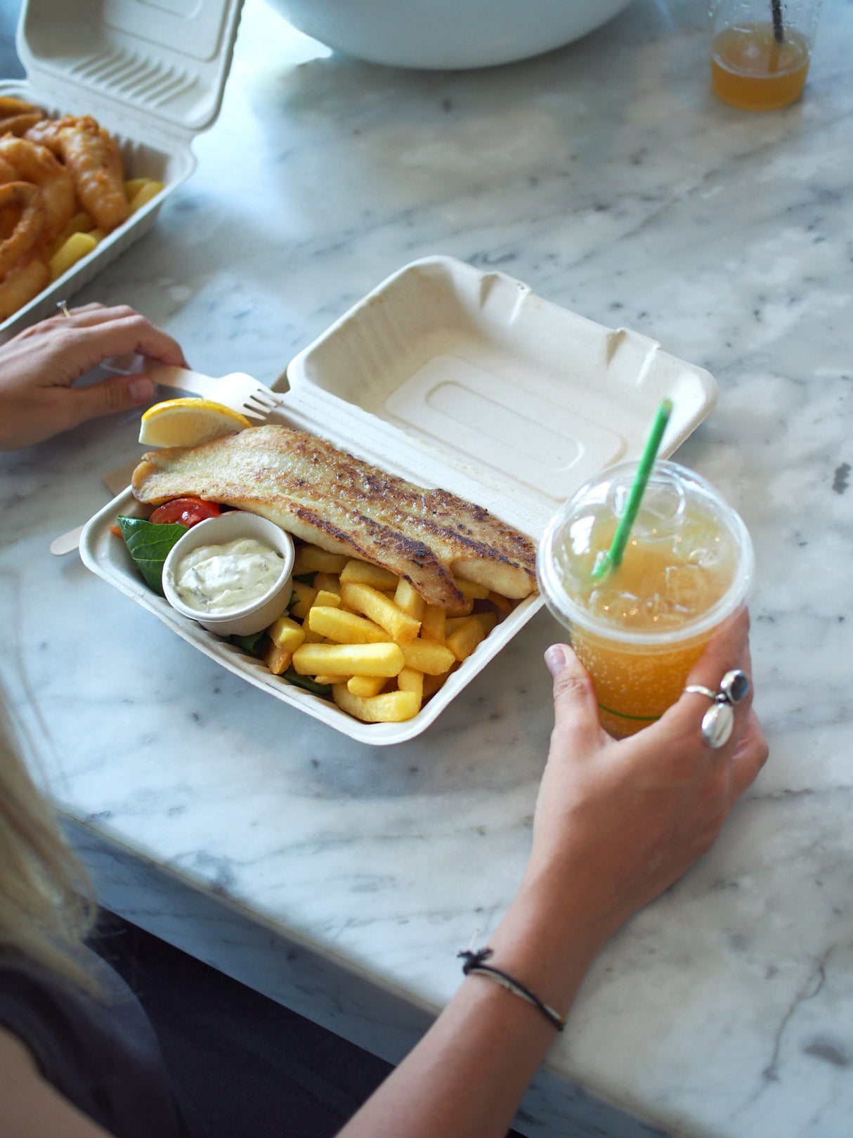 bagasse clamshell takeaway container size 9x6 containing fish and chips alongside a disposable sauce pot and a compostable clear takeaway cup with a paper straw in