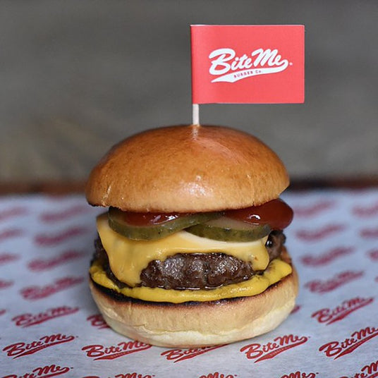 Beef burger with cheese and pickles, topped with a custom red burger flag with white lettering, served on custom printed greaseproof paper.