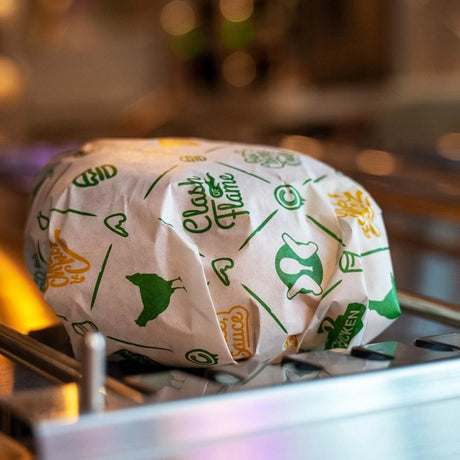 A burger wrapped in custom branded white and green greaseproof paper on a kitchen counter