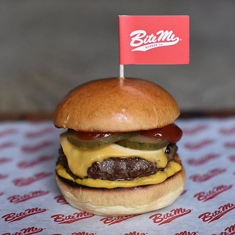Beef burger with cheese and pickles, topped with a custom red burger flag with white lettering, served on custom printed greaseproof paper.