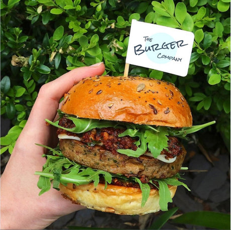 Hand holding a burger with 'The Burger Company' burger flag, against a green leafy background