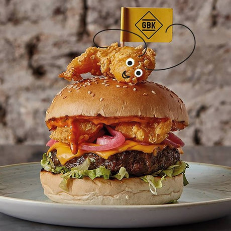 Hamburger with fried chicken on a bun, topped with a small character figure, on a plate with a GBK (gourmet burger kitchen) custom burger flag on top